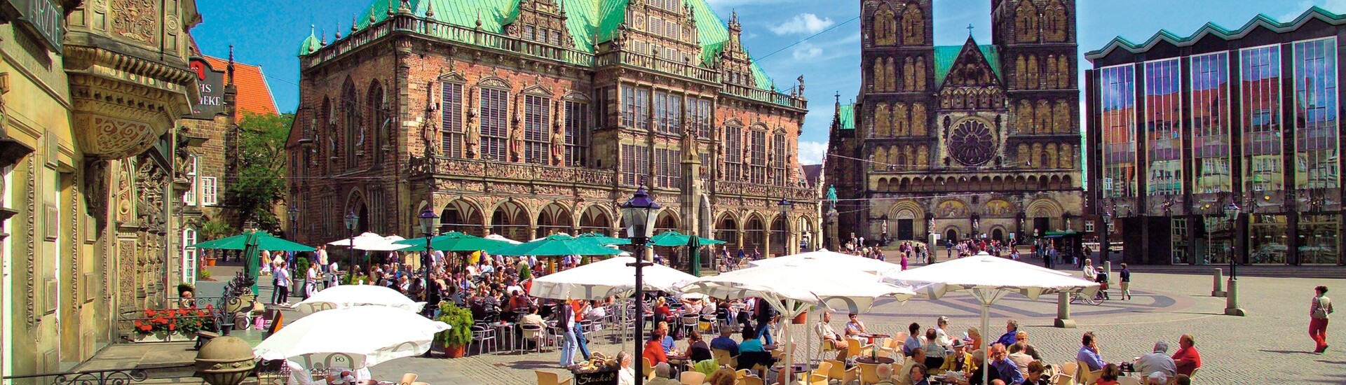 Belebter Platz mit historischem Gebäude, Dom und Café-Terrasse unter Sonnenschirmen bei blauem Himmel.