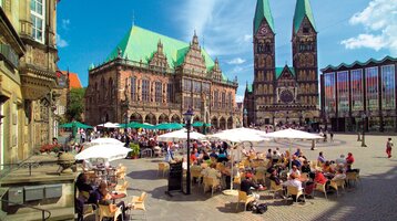 Belebter Platz mit historischem Gebäude, Dom und Café-Terrasse unter Sonnenschirmen bei blauem Himmel.