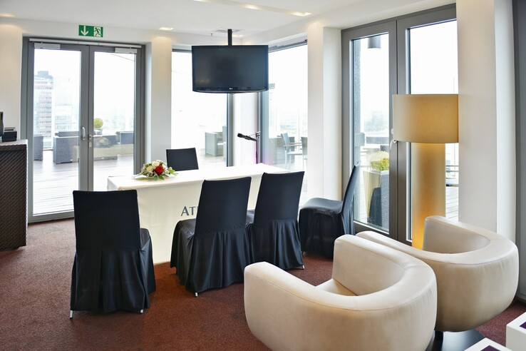 Moderner Konferenzraum im ATLANTIC Hotel Kiel mit Tisch, Stühlen, Blumenarrangement, TV und Blick auf die Terrasse.