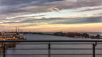 Blick von einem Balkon auf einen Fluss bei Sonnenuntergang, mit Stadtgebäuden und Wolken am Horizont.