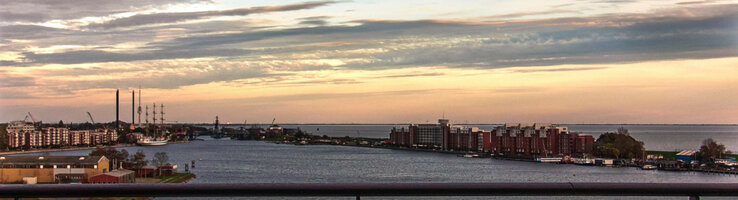 Blick von einem Balkon auf einen Fluss bei Sonnenuntergang, mit Stadtgebäuden und Wolken am Horizont.