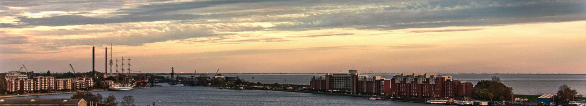 Angebot Best View | ATLANTIC Hotel Wilhelmshaven View from a balcony of a river at sunset, with city buildings and clouds on the horizon.