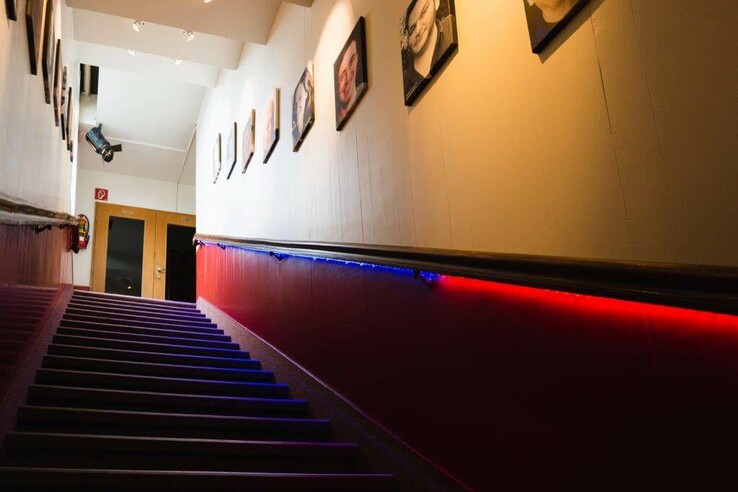 Staircase with red and blue lights, pictures on the walls, leading to a door in the background. Schnürschuh Theater Bremen