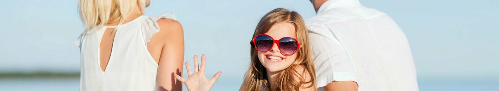 Familie am Strand in der Nähe Kiels Familie am Strand, sitzt auf einer Decke, lächelndes Kind mit roter Sonnenbrille, blauer Himmel im Hintergrund.