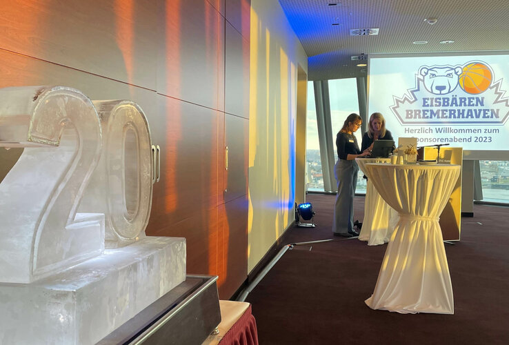Empfangsraum im ATLANTIC Hotel mit Eisbären Bremerhaven-Logo, Eisskulptur "20" und zwei Personen am Stehtisch.