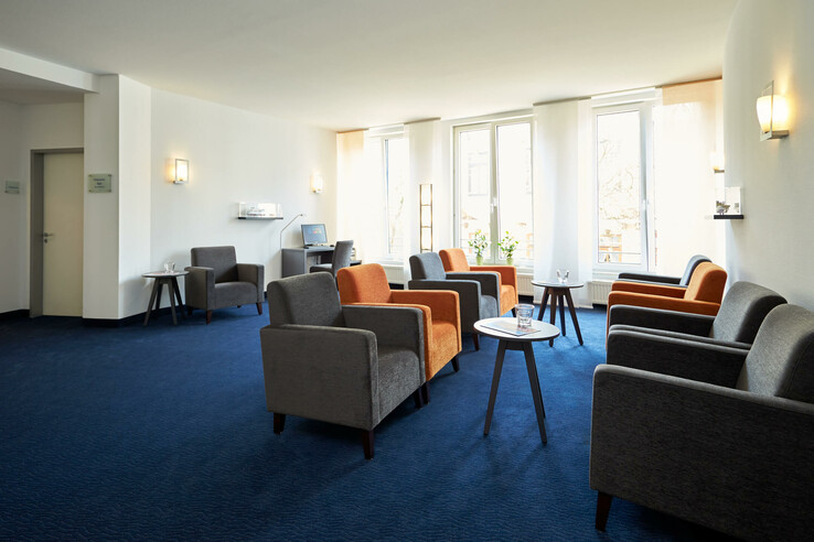 Heller Loungebereich mit grauen und orangefarbenen Sesseln, blauen Teppichboden und großen Fenstern im ATLANTIC Hotel Vegesack