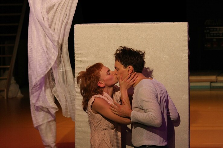 A couple kiss passionately in front of a white curtain in a warmly lit room. © bremer shakespeare company