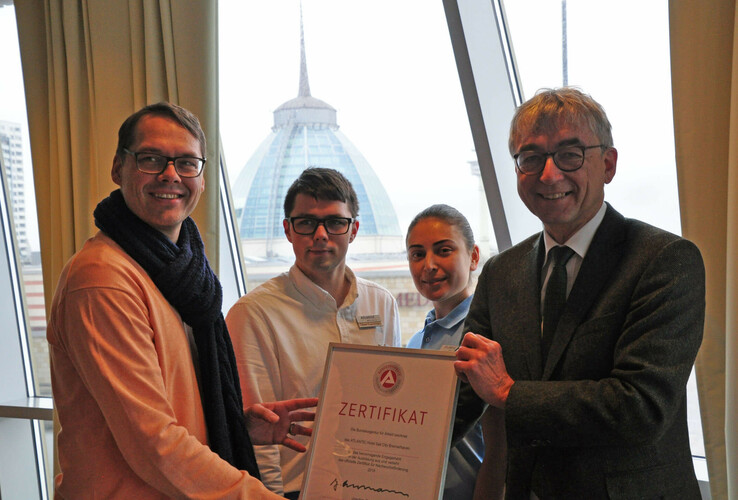 Vier Personen halten ein Zertifikat im ATLANTIC Hotel Sail City Bremerhaven mit dem Hotel im Hintergrund.