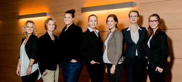 Eine Gruppe von sieben lächelnden Frauen in Business-Kleidung vor einer Holzwand im ATLANTIC Hotel Universum.