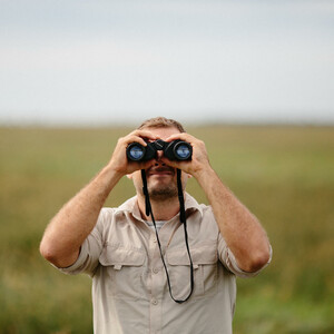 Man in beige shirt observes nature with binoculars in a wide, green landscape.