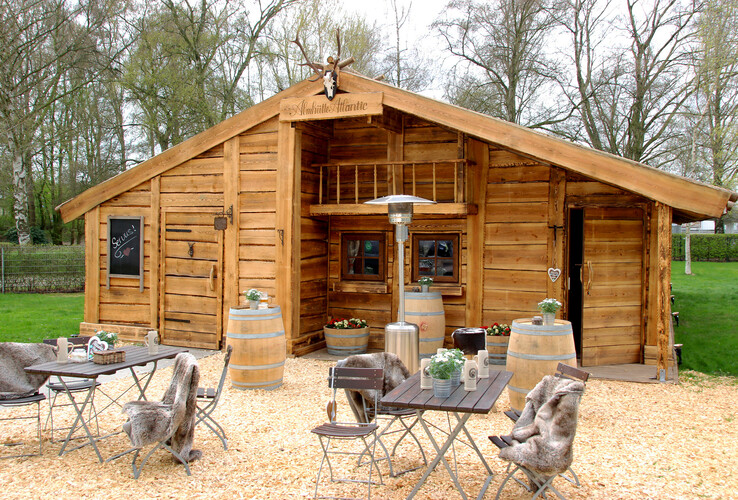 Gemütliche Holzhütte mit Außensitzbereich, Tischen, Stühlen und Pflanzen, umgeben von Bäumen und einem grünen Rasen.