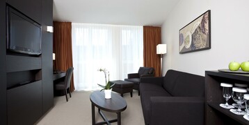 Modern hotel room with black sofa, coffee table, TV, desk, artwork on the wall and large windows.