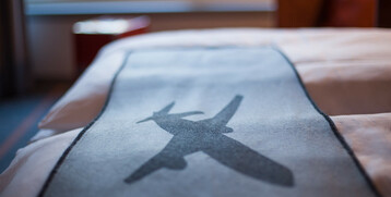 Bettdecke im ATLANTIC Hotel Airport Bremen mit Flugzeugmotiv, im Hintergrund verschwommene Fensteransicht.