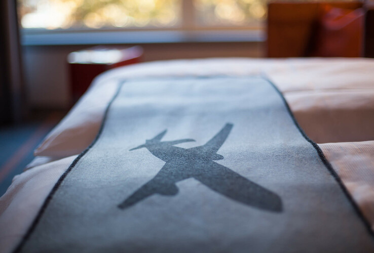 Bettdecke im ATLANTIC Hotel Airport Bremen mit Flugzeugmotiv, im Hintergrund verschwommene Fensteransicht.