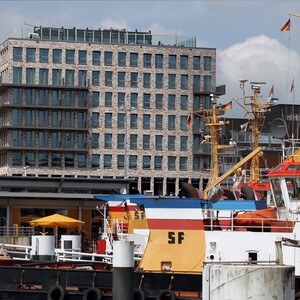 Modern hotel at the harbor with yellow and red ships in the foreground, blue sky in the background.