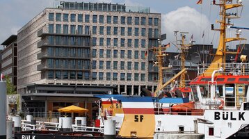 Modernes Hotel am Hafen mit gelben und roten Schiffen im Vordergrund, blauem Himmel im Hintergrund.