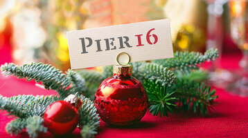 Weihnachtlicher Tannenzweig und Weihnachtskugel halten ein "PIER16" Schild