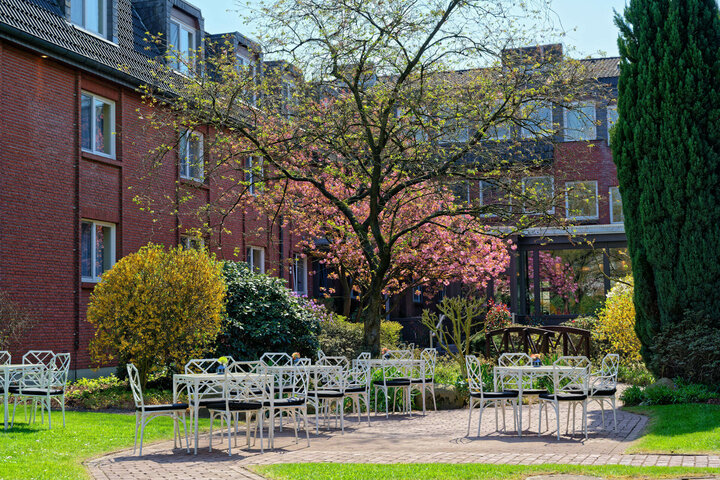 Garden view with blossoming tree, white garden furniture and red brick building at the ATLANTIC Hotel Landgut Horn Bremen.