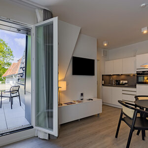 Essbereich in der Kategorie Appartement Plus in der ATLANTIC Dependance | ATLANTIC Grand Hotel Travemünde Helles Hotelzimmer mit Küchenzeile, Esstisch und Zugang zu einem Balkon mit Sitzbereich und Blick auf grüne Bäume.