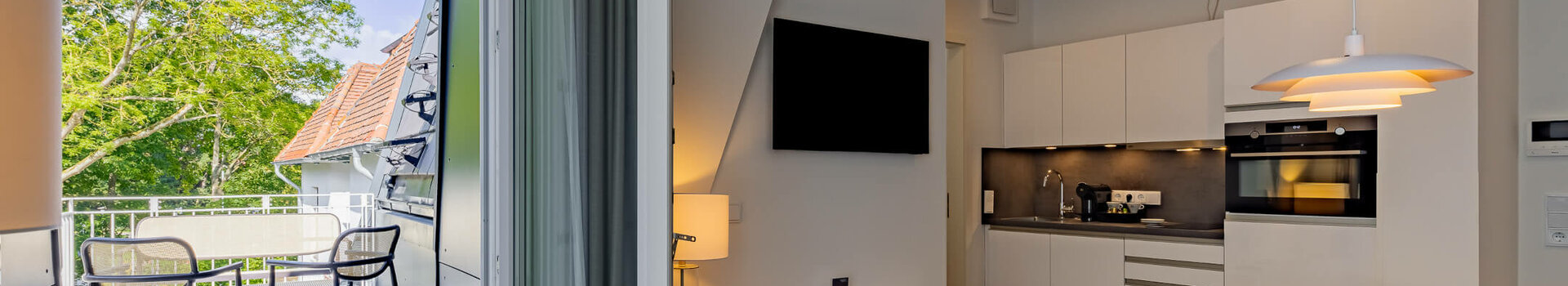 Essbereich in der Kategorie Appartement Plus in der ATLANTIC Dependance | ATLANTIC Grand Hotel Travemünde Helles Hotelzimmer mit Küchenzeile, Esstisch und Zugang zu einem Balkon mit Sitzbereich und Blick auf grüne Bäume.