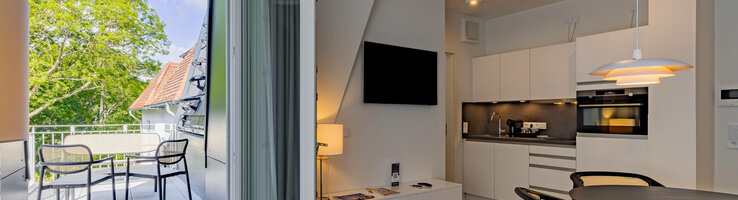 Essbereich in der Kategorie Appartement Plus in der ATLANTIC Dependance | ATLANTIC Grand Hotel Travemünde Helles Hotelzimmer mit Küchenzeile, Esstisch und Zugang zu einem Balkon mit Sitzbereich und Blick auf grüne Bäume.