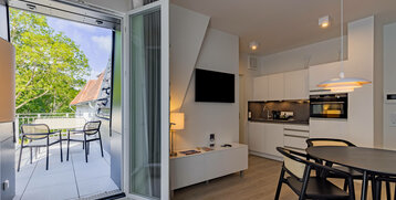 Bright hotel room with kitchenette, dining table and access to a balcony with seating area and view of green trees.