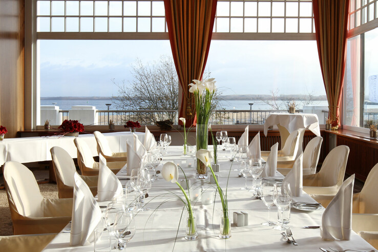 Elegantes Restaurant mit Meerblick, gedeckten Tischen, weißen Tischdecken und Blumenarrangement in einem hellen Raum.