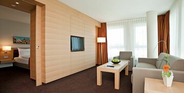 Helles Hotelzimmer mit Sofa, Sessel, Couchtisch, Flachbild-TV und großen Fenstern mit Vorhängen im ATLANTIC Hotel Kiel.