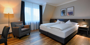 Helles Hotelzimmer mit Doppelbett, Sessel, Lampe und Bildern an der Wand, ATLANTIC Hotel Landgut Horn Bremen.