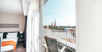 Helles Hotelzimmer mit Balkon, Blick auf Lübecker Kirchtürme, modernes Design, gemütliches Bett mit orangefarbener Decke.