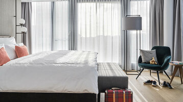 Innenansicht Zimmer Studio Helles Hotelzimmer mit großem Bett, roten Kissen, Sessel, Stehlampe und Koffer auf Holzfußboden.