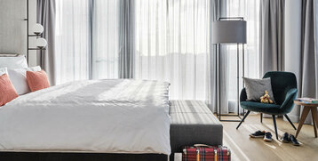 Helles Hotelzimmer mit großem Bett, roten Kissen, Sessel, Stehlampe und Koffer auf Holzfußboden.