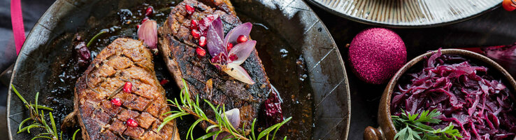 Gebratene Entenbrust in Pfanne mit Granatapfel, Rosmarin und Rotkohl, festlich dekoriert mit roten Kugeln.