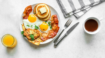 Ein Frühstücksteller mit Pancakes, Speck, Spiegeleiern und Kartoffelrösti, dazu Kaffee und Orangensaft