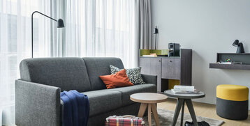 Modernes Hotelzimmer mit grauem Sofa, Couchtischen, Kaffeemaschine und dekorativen Kissen. Helle, einladende Atmosphäre.