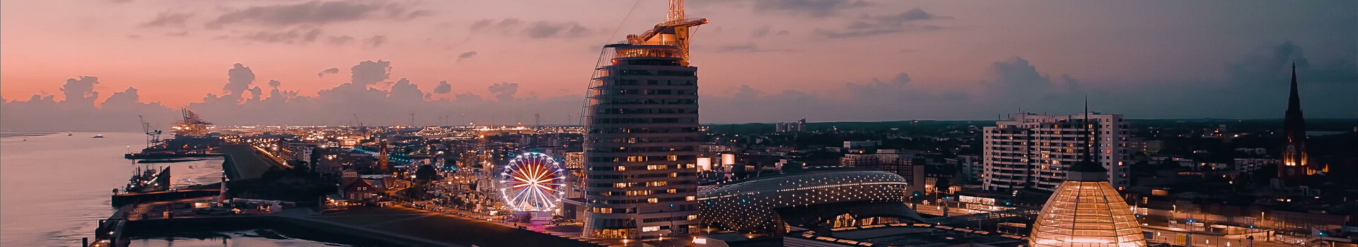 EDM meets Classic Abendliche Skyline von Bremerhaven mit ATLANTIC Hotel Sail City und beleuchtetem Riesenrad am Wasser.