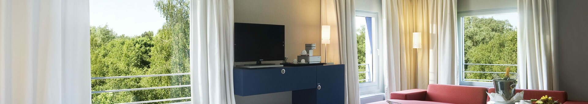 Interior view of a Studio in the ATLANTIC Hotel Universum with a spacious living area Modern hotel room with two beds, red sofa, large window and view of green trees in the ATLANTIC Hotel Universum.