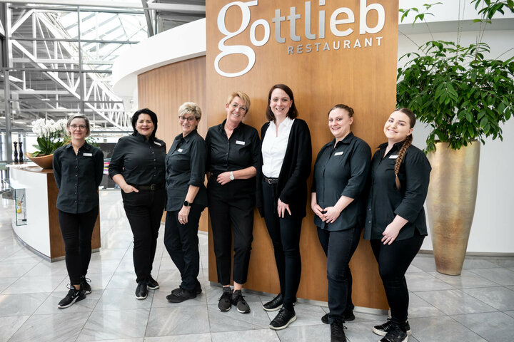 Mitarbeiter des Restaurants gottlieb im Mercedes-Benz Kundencenter Sieben Restaurantmitarbeiterinnen in schwarzer Kleidung lächeln vor einem Holzschild mit "Gottlieb Restaurant".