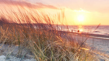 Sonnenuntergang am Strand von Travemünde, mit Gras im Vordergrund und Personen am Wasser.