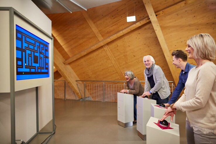 Vier Personen spielen gemeinsam ein Retro-Videospiel auf einem großen Bildschirm in einem modernen Raum mit Holzwänden.