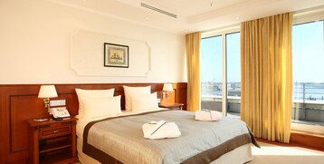 Schlafzimmer der Kaiser Wilhelm Suite mit großem Bett und Blick auf den Hafen durch große Fenster im ATLANTIC Hotel Wilhelmshaven