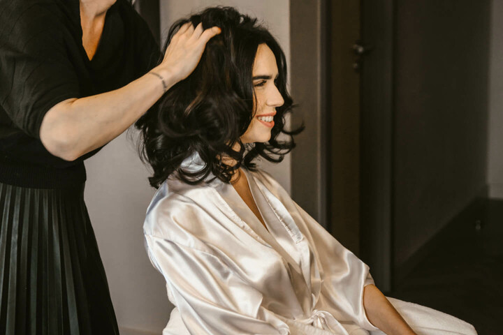 Frau im Bademantel sitzt lächelnd, während eine Stylistin ihr die Haare macht, in einem eleganten Hotelzimmer.