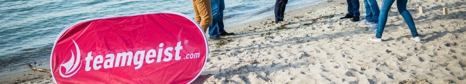 Menschen spielen ein Teamspiel am Strand, nahe dem ATLANTIC Grand Hotel Travemünde, mit einem "teamgeist.com"-Banner.