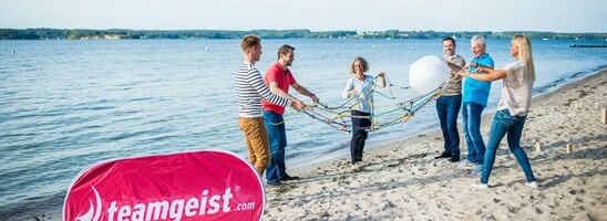 Menschen spielen ein Teamspiel am Strand, nahe dem ATLANTIC Grand Hotel Travemünde, mit einem "teamgeist.com"-Banner.
