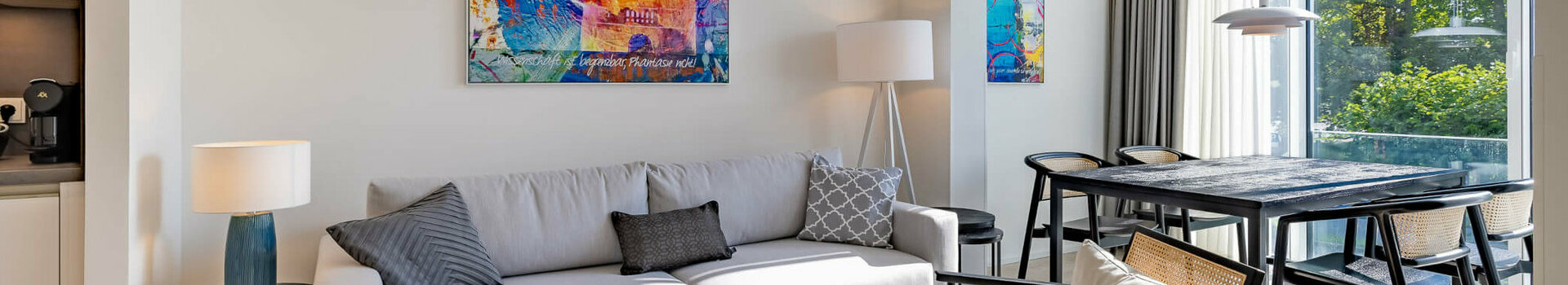 Wohnbereich in der Kategorie Studio Plus in der ATLANTIC Dependance | ATLANTIC Grand Hotel Travemünde Modernes Hotelzimmer mit grauem Sofa, buntem Kunstwerk, Esstisch und großen Fenstern mit Blick ins Grüne.