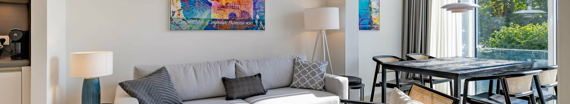 Living area in the Studio Plus category in the ATLANTIC Dependance | ATLANTIC Grand Hotel Travemünde Modern hotel room with gray sofa, colorful artwork, dining table and large windows overlooking the greenery.