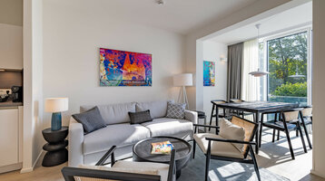 Modernes Hotelzimmer mit grauem Sofa, buntem Kunstwerk, Esstisch und großen Fenstern mit Blick ins Grüne.