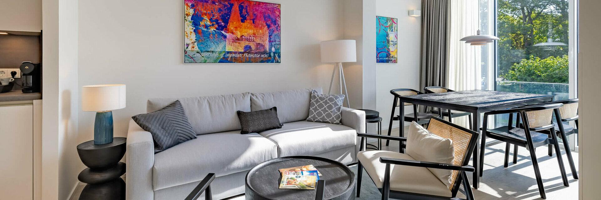 Modern hotel room with gray sofa, colorful artwork, dining table and large windows overlooking the greenery.