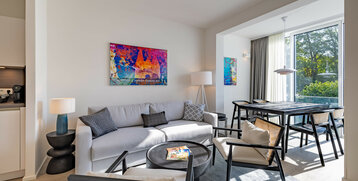 Modern hotel room with gray sofa, colorful artwork, dining table and large windows overlooking the greenery.