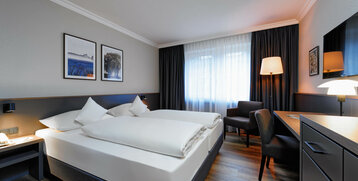 Modernes Hotelzimmer mit zwei Einzelbetten, Schreibtisch, Sessel, Stehlampe und Bildern an der Wand.